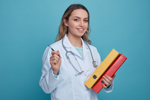 Smiling young female doctor wearing medical robe and stethoscope around neck holding pen and folders 