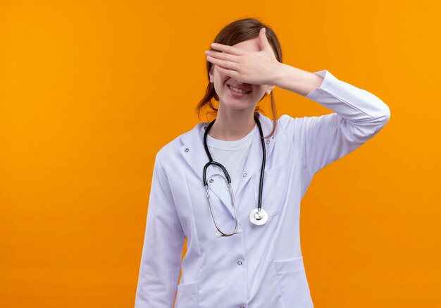 Улыбающаяся молодая женщина-врач в медицинском халате и стетоскопе, закрывающая глаза рукой на изолированной оранжевой стене с копией пространства