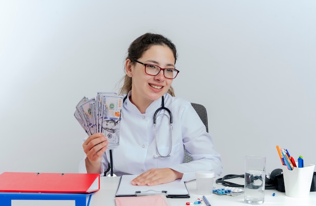 Smiling young female doctor wearing medical robe and stethoscope and glasses sitting at desk with medical tools holding money looking at side isolated
