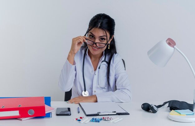 Smiling young female doctor wearing medical robe and stethoscope and glasses sitting at desk with medical tools looking putting hand on desk grabbing glasses isolated