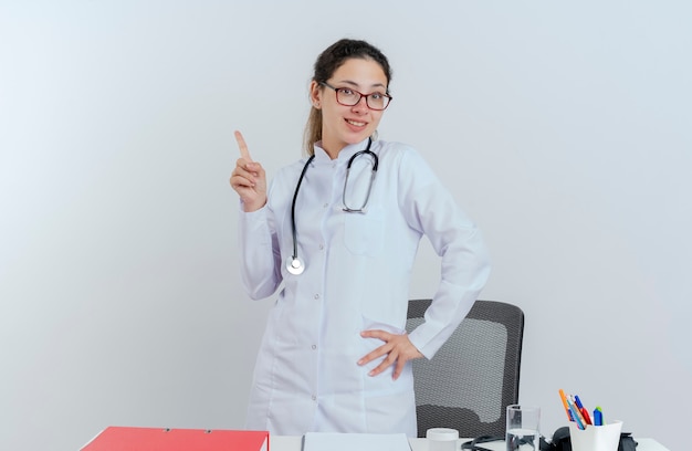 Smiling young female doctor wearing medical robe and stethoscope and glasses standing behind desk with medical tools looking putting hand on waist raising finger isolated