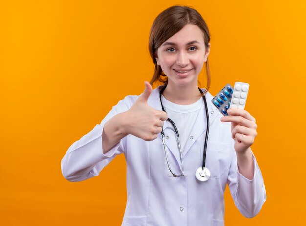 Smiling young female doctor wearing medical robe and stethoscope holding medical drugs and showing thumb up on isolated orange wall