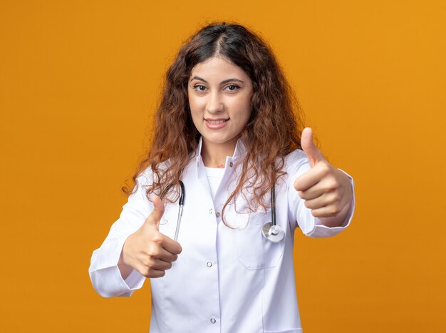 オレンジ色の壁に分離された親指を見せて正面を見て医療ローブと聴診器を身に着けている若い女性医師の笑顔