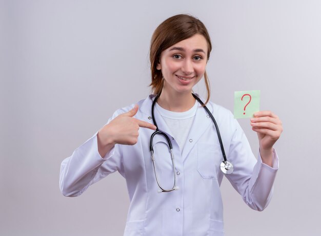 Smiling young female doctor wearing medical robe and stethoscope and ponting at question mark written on paper note on isolated white wall