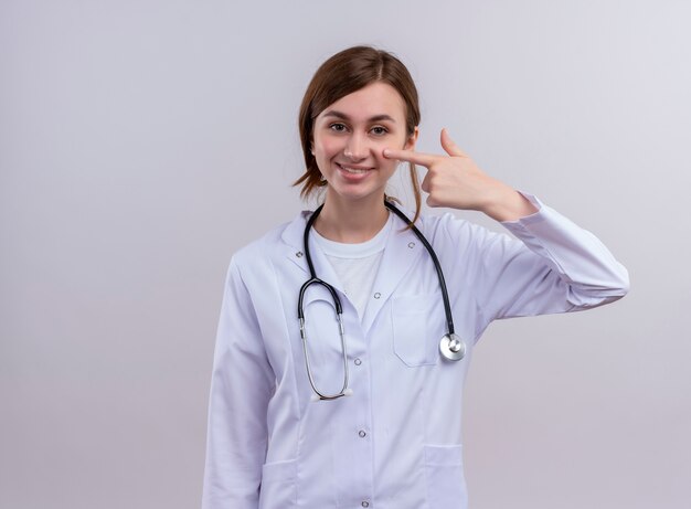 의료 가운과 청진 기 복사 공간이 격리 된 흰 벽에 뺨에 손가락을 넣어 입고 웃는 젊은 여성 의사