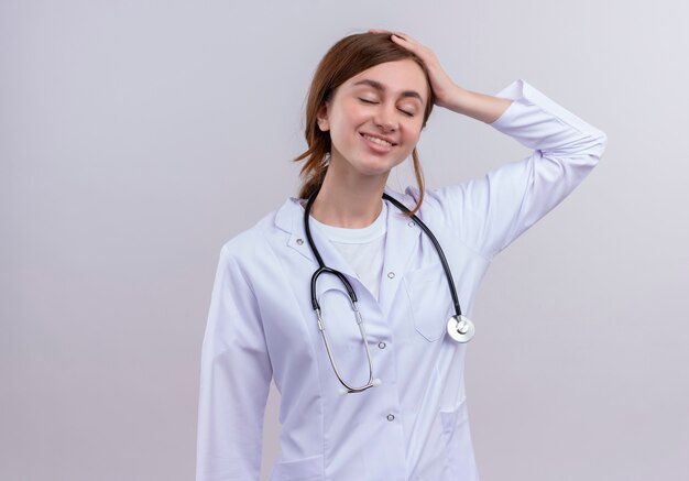 Улыбающаяся молодая женщина-врач в медицинском халате и стетоскопе, положив руку на голову на изолированной белой стене с копией пространства
