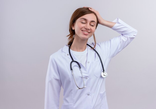 Smiling young female doctor wearing medical robe and stethoscope and putting hand on head on isolated white wall with copy space