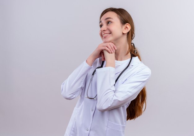 Smiling young female doctor wearing medical robe and stethoscope putting hands under chin on isolated white wall with copy space