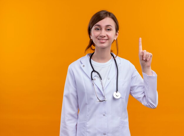 의료 가운 및 복사 공간이 격리 된 오렌지 벽에 손가락을 올리는 청진기를 입고 웃는 젊은 여성 의사