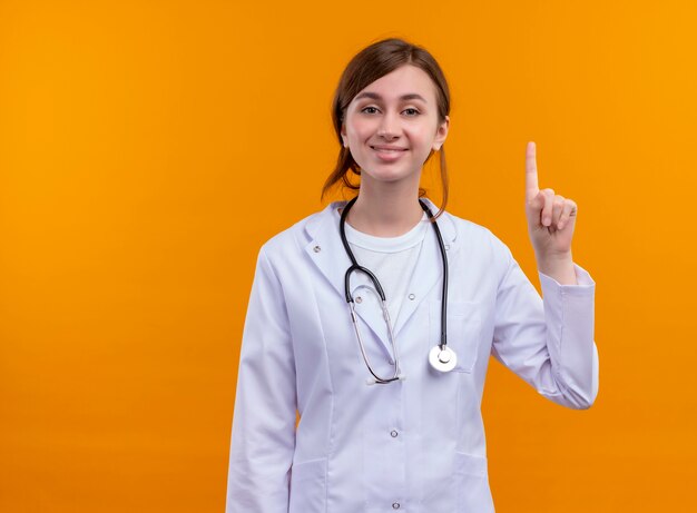 Smiling young female doctor wearing medical robe and stethoscope raising finger on isolated orange wall with copy space