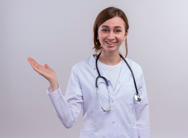 의료 가운과 청진기를 착용하고 격리 된 흰 벽에 빈 손을 보여주는 웃는 젊은 여성 의사