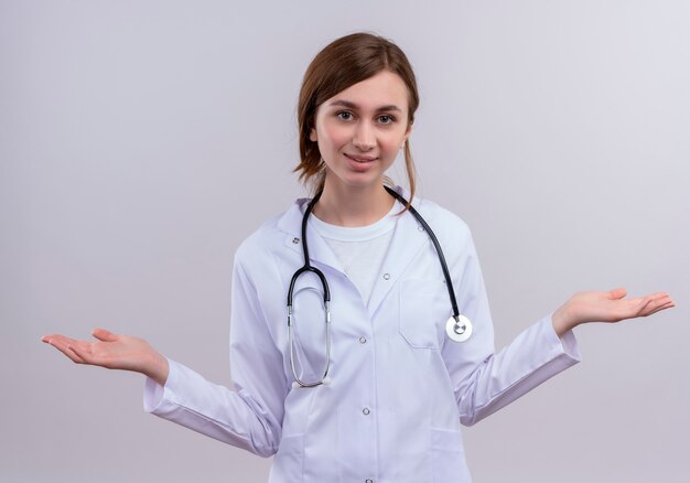 격리 된 흰 벽에 빈 손을 보여주는 의료 가운과 청진기를 입고 웃는 젊은 여성 의사