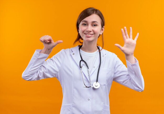 孤立したオレンジ色の壁に6を示す医療ローブと聴診器を身に着けている若い女性医師の笑顔