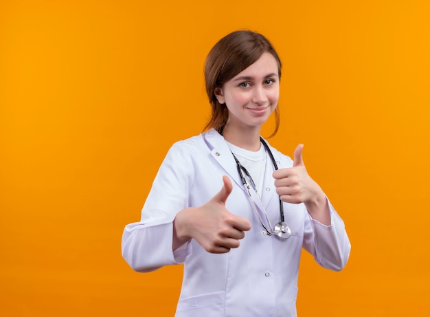 コピースペースと孤立したオレンジ色の壁に親指を示す医療ローブと聴診器を身に着けている若い女性医師の笑顔