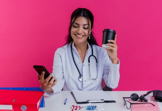 분홍색 벽에 고립 된 전화를 찾고 커피 컵과 휴대 전화를 들고 의료 도구와 책상에 앉아 의료 가운과 청진기를 입고 웃는 젊은 여성 의사