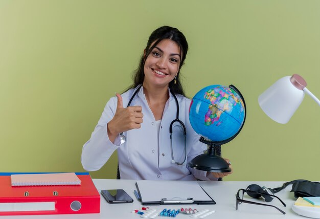 Smiling young female doctor wearing medical robe and stethoscope sitting at desk with medical tools holding globe looking showing thumb up isolated
