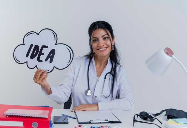 Smiling young female doctor wearing medical robe and stethoscope sitting at desk with medical tools holding idea bubble looking putting hand on desk isolated
