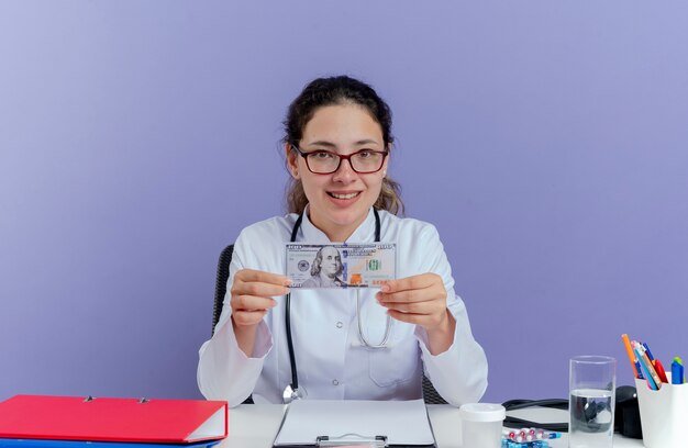 Smiling young female doctor wearing medical robe and stethoscope sitting at desk with medical tools holding money looking isolated