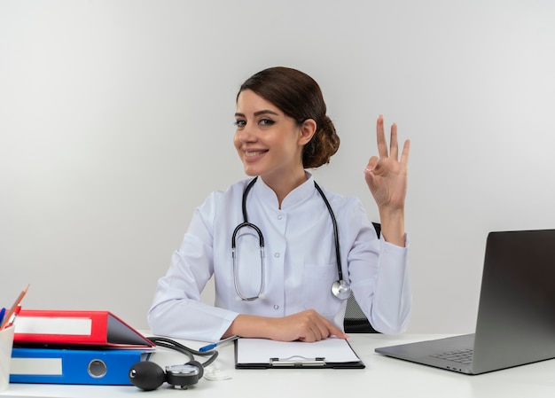 Smiling young female doctor wearing medical robe and stethoscope sitting at desk with medical tools and laptop doing ok sign isolated on white wall