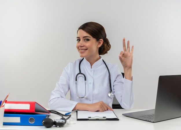 의료 가운과 청진기를 입고 의료 도구와 노트북 흰 벽에 고립 된 확인 서명을 하 고 책상에 앉아 웃는 젊은 여성 의사
