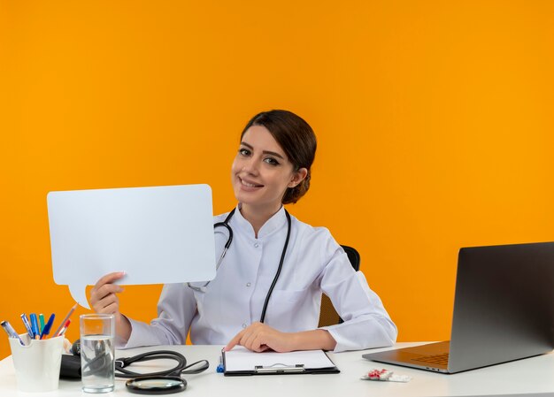 Smiling young female doctor wearing medical robe and stethoscope sitting at desk with medical tools and laptop holding chat bubble isolated on yellow wall