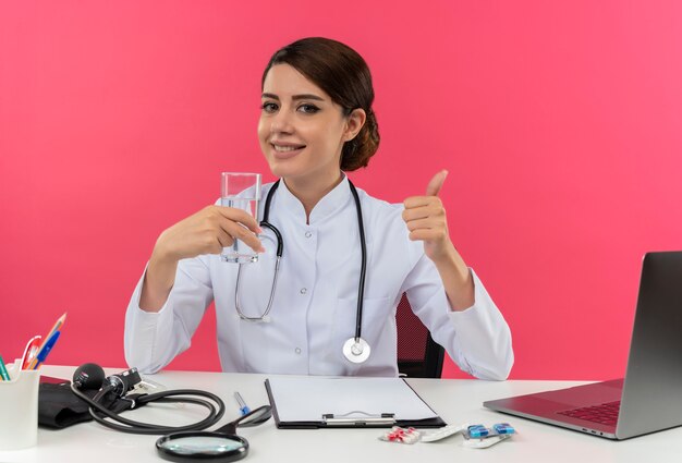 Улыбающаяся молодая женщина-врач в медицинском халате и стетоскопе сидит за столом с медицинскими инструментами и ноутбуком, держа стакан воды и показывая большой палец вверх