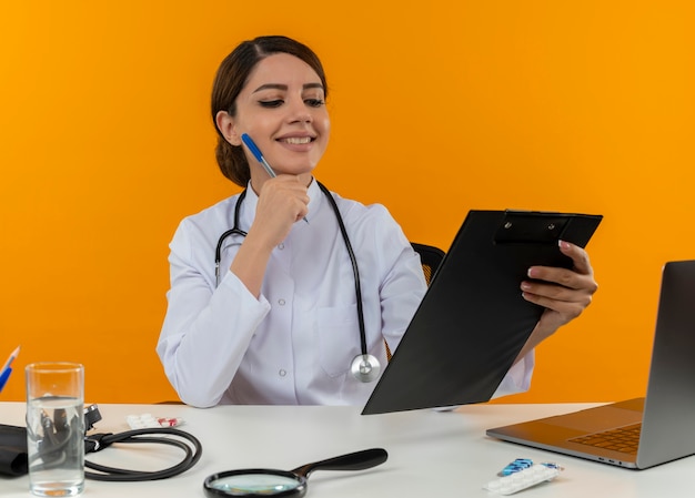 Smiling young female doctor wearing medical robe and stethoscope sitting at desk with medical tools and laptop holding and looking at clipboard touching face with pen isolated on yellow wall