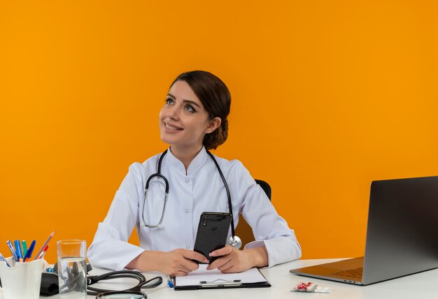 Smiling young female doctor wearing medical robe and stethoscope sitting at desk with medical tools and laptop holding mobile phone looking at side isolated on yellow wall