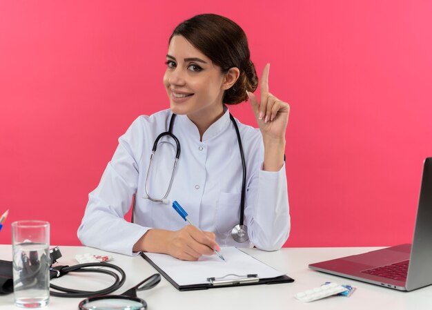 Улыбающаяся молодая женщина-врач в медицинском халате и стетоскопе сидит за столом с медицинскими инструментами и ноутбуком, держа ручку и поднимая палец, изолированные на розовой стене