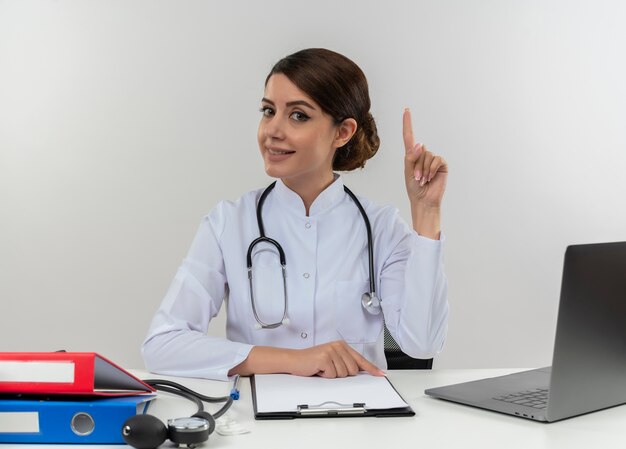 Smiling young female doctor wearing medical robe and stethoscope sitting at desk with medical tools and laptop pointing up isolated on white wall