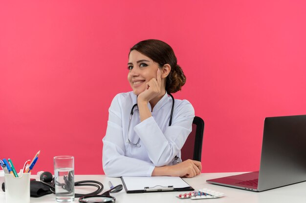 의료용 가운과 청진기를 착용하고 의료 도구와 노트북이 책상에 손을 얹고 턱에 똑바로 분홍색 벽에 고립 된 찾고 책상에 앉아 웃는 젊은 여성 의사