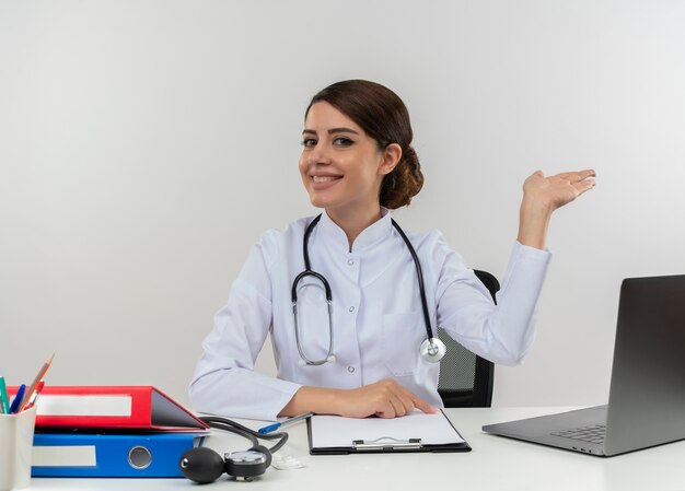 Smiling young female doctor wearing medical robe and stethoscope sitting at desk with medical tools and laptop showing empty hand isolated on white wall