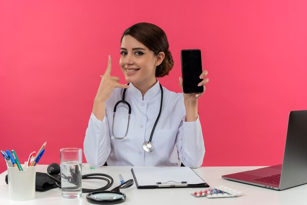 Smiling young female doctor wearing medical robe and stethoscope sitting at desk with medical tools and laptop showing mobile phone doing hang loose gesture 