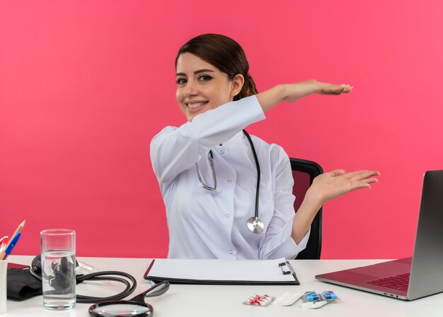 Smiling young female doctor wearing medical robe and stethoscope sitting at desk with medical tools and laptop showing size isolated on pink wall