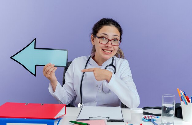 Smiling young female doctor wearing medical robe and stethoscope sitting at desk with medical tools looking holding arrow mark pointing at side isolated