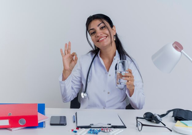 Smiling young female doctor wearing medical robe and stethoscope sitting at desk with medical tools looking holding glass of water doing ok sign isolated