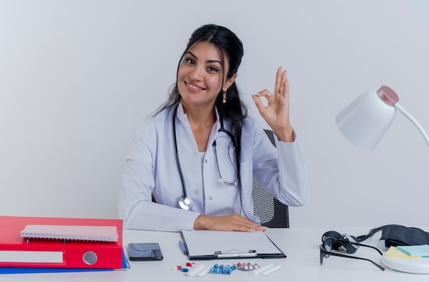 Foto gratuita sorridente giovane medico femminile che indossa abito medico e stetoscopio seduto alla scrivania con strumenti medici cercando di mettere la mano sulla scrivania facendo segno ok isolato