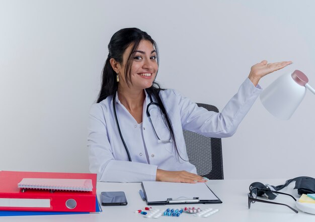 医療用ローブと聴診器を身に着けている笑顔の若い女性医師が机の上に手を置いて机の上に手を置いて机に座っている
