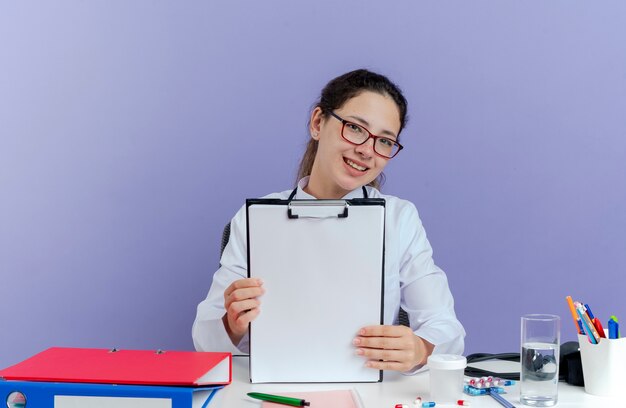 Sorridente giovane medico femminile che indossa abito medico e stetoscopio seduto alla scrivania con strumenti medici cercando mostrando appunti isolati