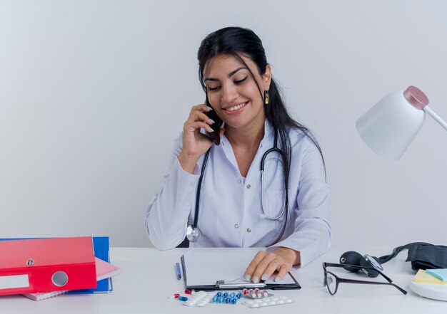 Smiling young female doctor wearing medical robe and stethoscope sitting at desk with medical tools putting hand on clipboard talking on phone looking down isolated