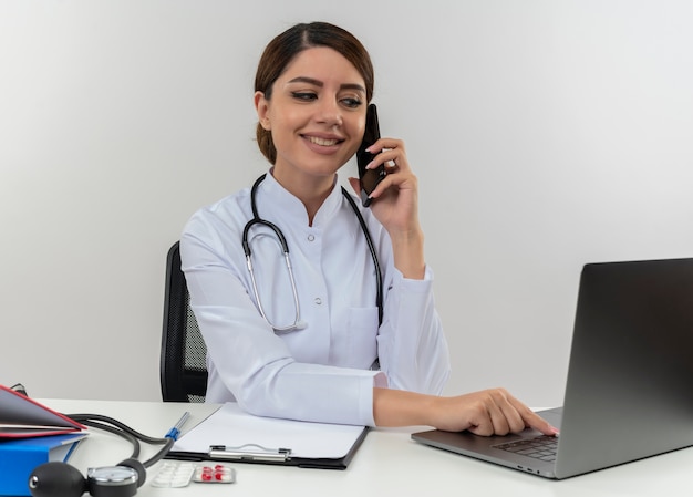 Smiling young female doctor wearing medical robe and stethoscope sitting at desk with medical tools using and looking at laptop talking on phone isolated on white wall