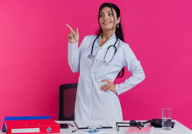 Smiling young female doctor wearing medical robe and stethoscope standing behind desk with medical tools keeping hand on waist looking and pointing at side isolated