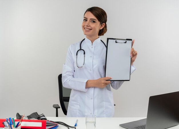 Smiling young female doctor wearing medical robe and stethoscope standing behind desk with medical tools and laptop holding clipboard isolated on white wall