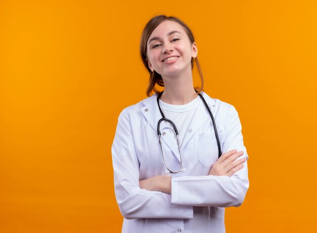 コピースペースと孤立したオレンジ色の壁に閉じた姿勢で立っている医療ローブと聴診器を身に着けている若い女性医師の笑顔