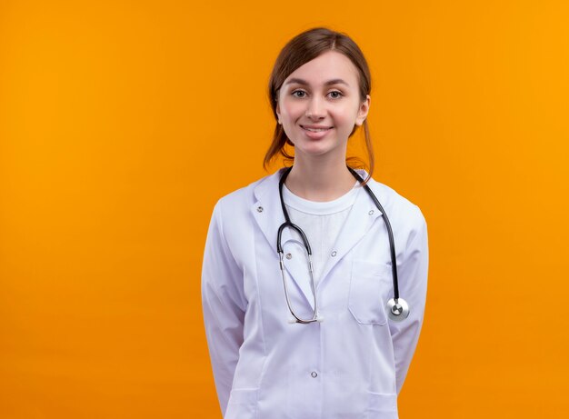 Smiling young female doctor wearing medical robe and stethoscope standing with hands behind her back on isolated orange wall with copy space