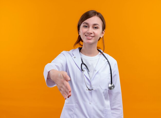 Улыбающаяся молодая женщина-врач в медицинском халате и стетоскопе протягивает руку на изолированной оранжевой стене с копией пространства