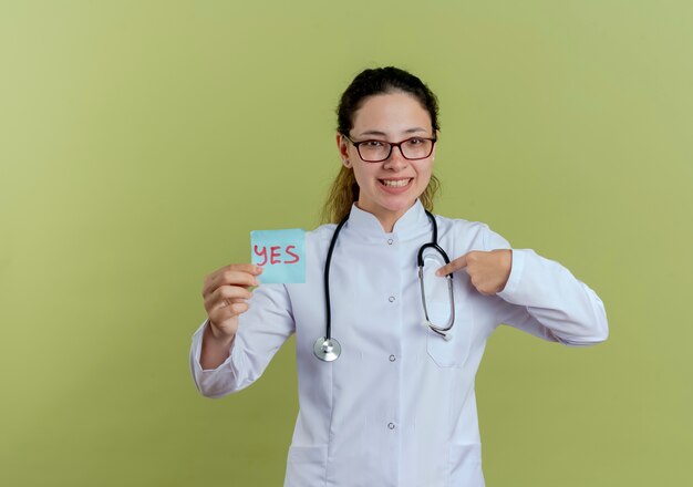 Smiling young female doctor wearing medical robe and stethoscope with glasses holding paper note points at herself isolated on olive green wall