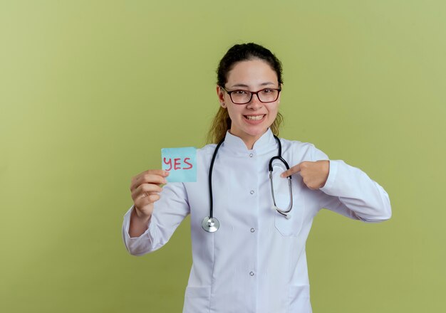 Улыбающаяся молодая женщина-врач в медицинском халате и стетоскопе в очках, держащая бумажные записки, указывает на себя, изолированную на оливково-зеленой стене