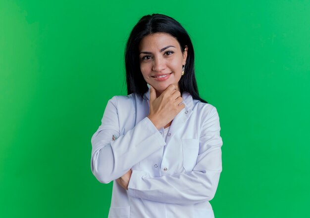 Smiling young female doctor wearing medical robe touching chin isolated on green wall with copy space