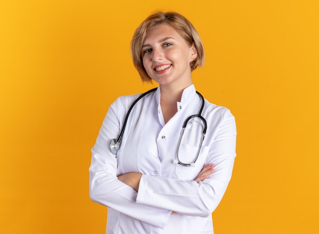 Smiling young female doctor wearing medical robe with stethoscope crossing hands isolated on orange background